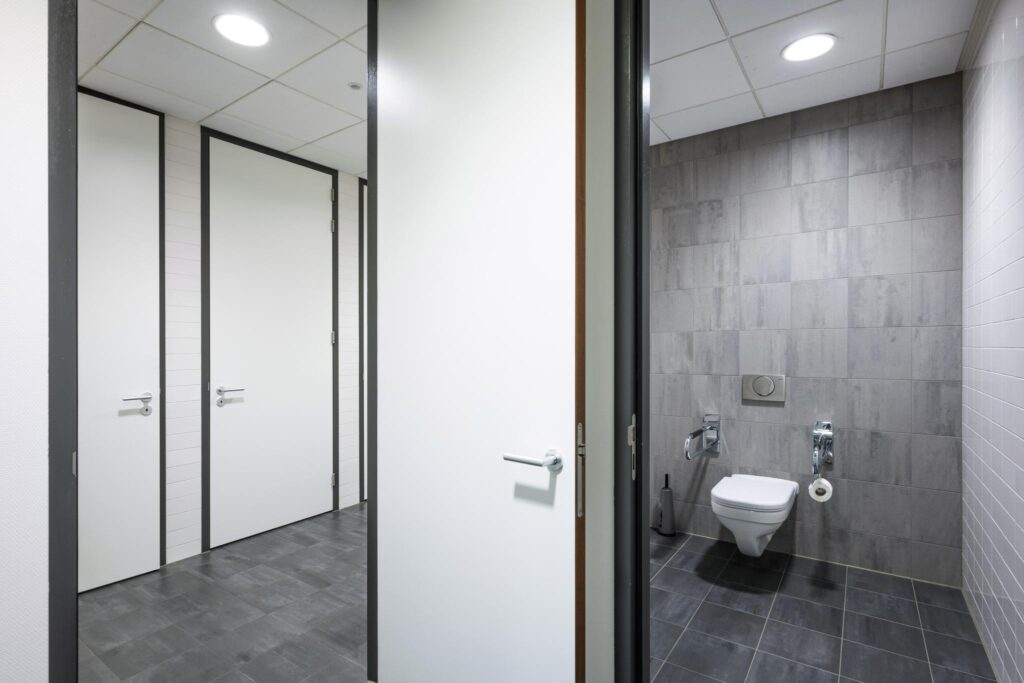 Publieke toiletruimte met grijze vloertegels en een openstaande deur naar een modern ingericht toilet in een gebouw aan de Laan van de Helende Meesters.